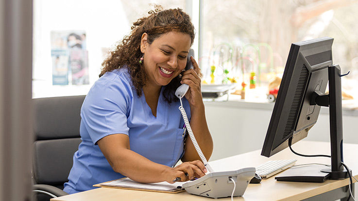 A nurse smiles as she sits in her office in front of a computer and speaks on the phone